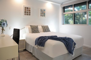 Ein Schlafzimmer mit Bett, Nachttisch, Stuhl, Fenster mit Blick auf Bäume und zwei gerahmte Bilder an der Wand.