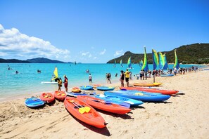 A beach with multiple kayaks and canoes lined up on the sand, a clear blue sea, and a hilly landscape in the background.