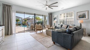 A spacious living room with a large grey sofa, a ceiling fan, and a view of a pool and palm trees outside.