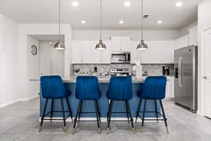 A modern kitchen with white cabinets, a stainless steel refrigerator, a microwave, and a bar area with blue bar stools. - Windsor Oasis | Kids Bedrooms | Private Pool | Townhome (Davenport)
