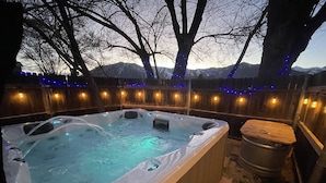Ein Whirlpool mit einer Holzbank, umgeben von einem Holzzaun mit Lichterketten, vor einer Kulisse aus Bäumen und Bergen.