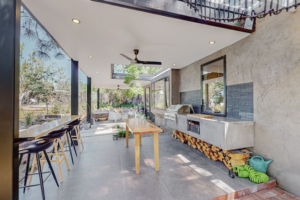 An outdoor dining area with a long wooden table and black bar stools, a barbecue grill, and a view of trees and a fence.