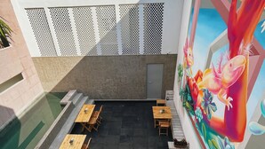 Un patio al aire libre con mesas y bancos de madera, una piscina y un mural con peces y flores en la pared.