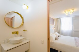 A hotel room with a round mirror, a white sink, a bed with white bedding, and a chandelier.