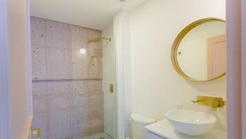 Un baño con ducha envuelta en vidrio, inodoro blanco y un lavamanos grande de color blanco con grifo dorado.