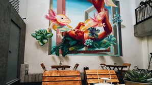 Un comedor con mesas y sillas de madera, un mural con una mujer y peces, y suculentas en la pared.