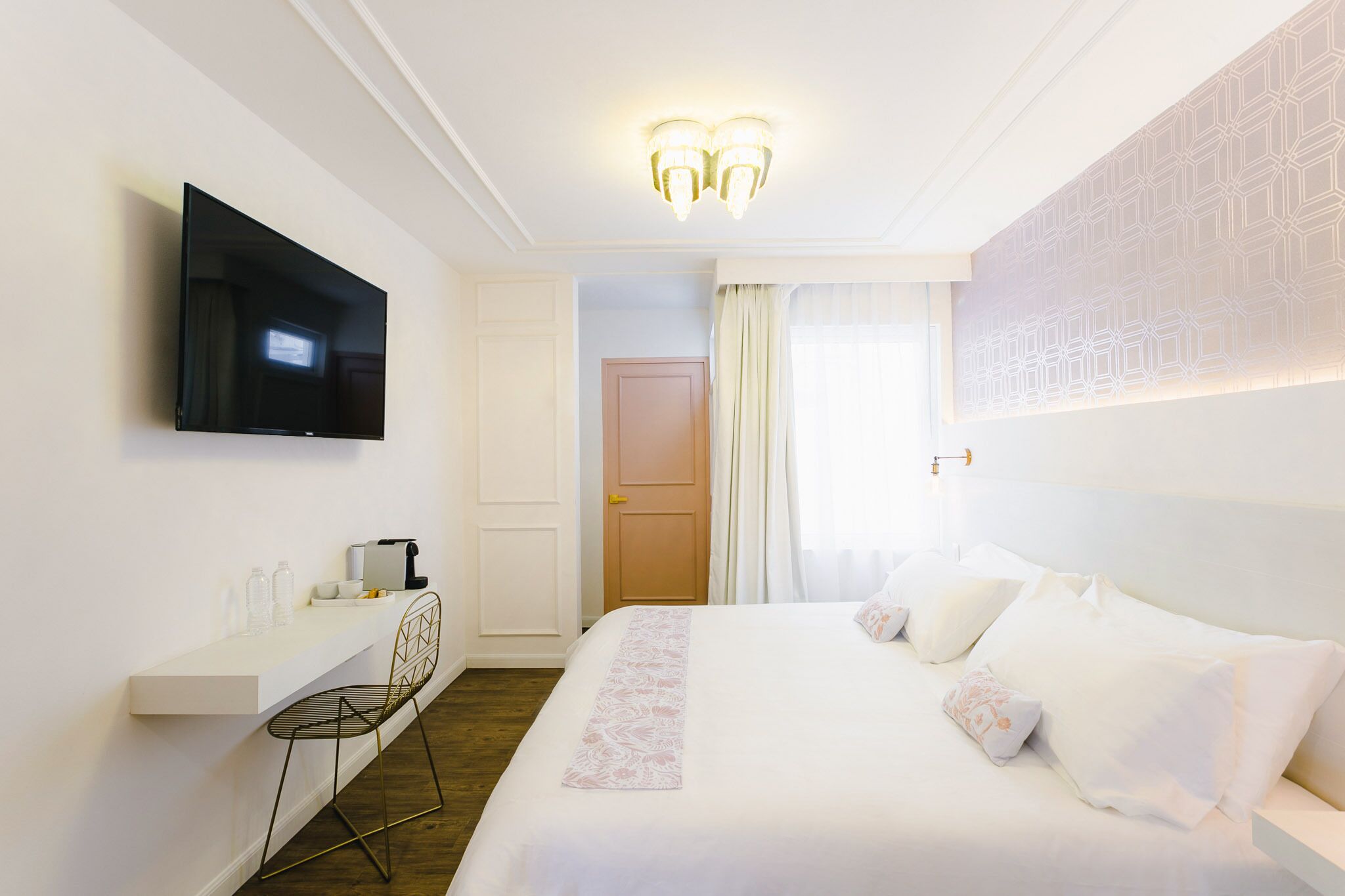 A hotel room with a bed, a TV mounted on the wall, a desk with a chair, and a window with curtains.