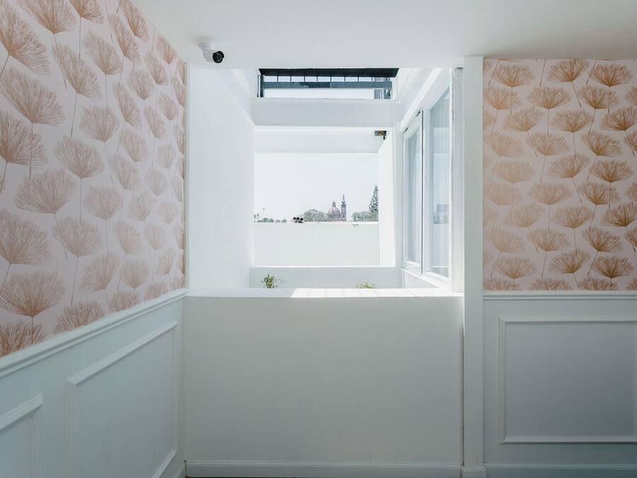 Una habitación con paredes blancas, una ventana que da a un edificio y papel tapiz estampado.