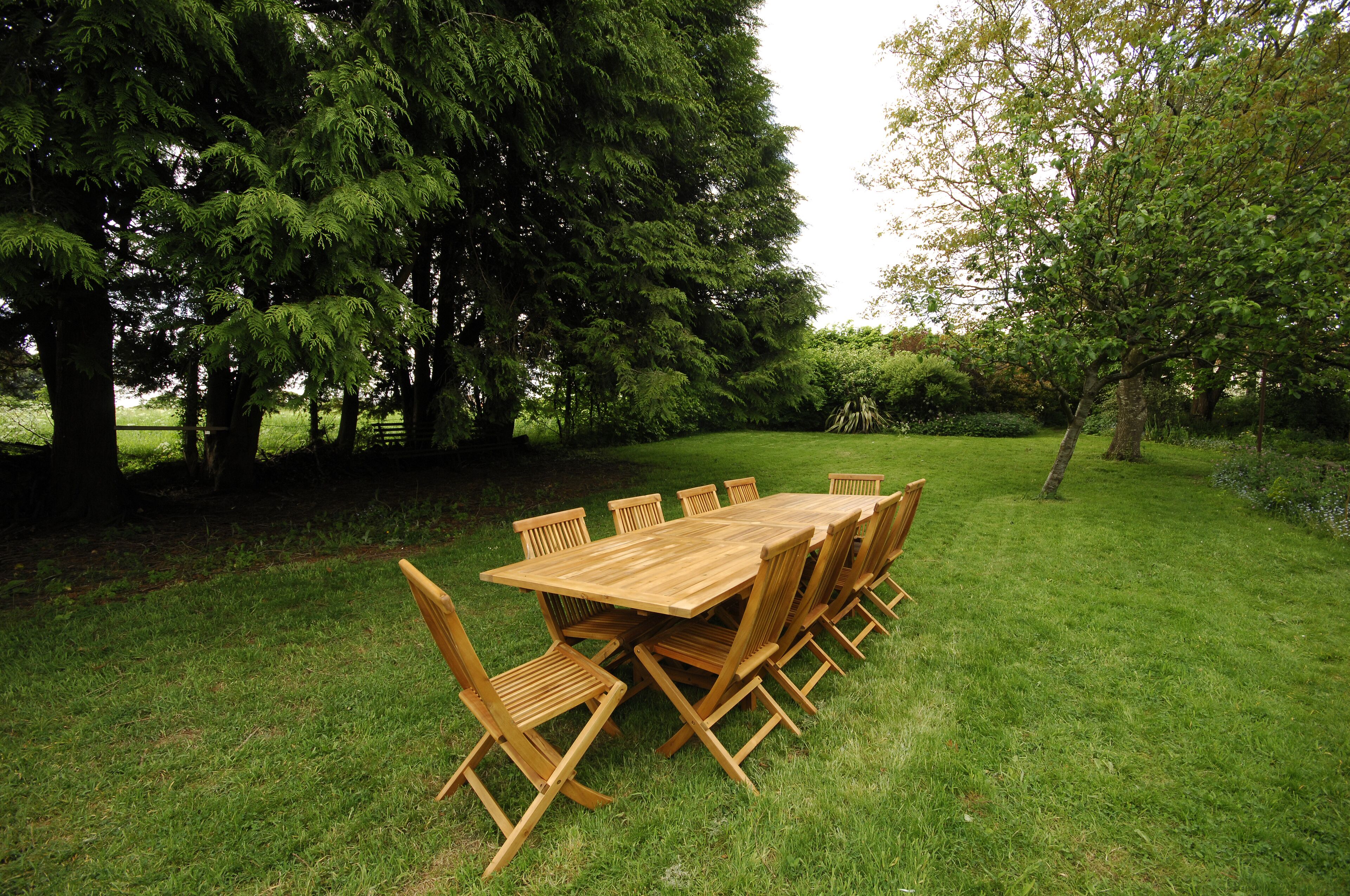 Une table à manger en bois, avec des chaises, est placée à l’extérieur, sur une pelouse verdoyante, entourée d’arbres.