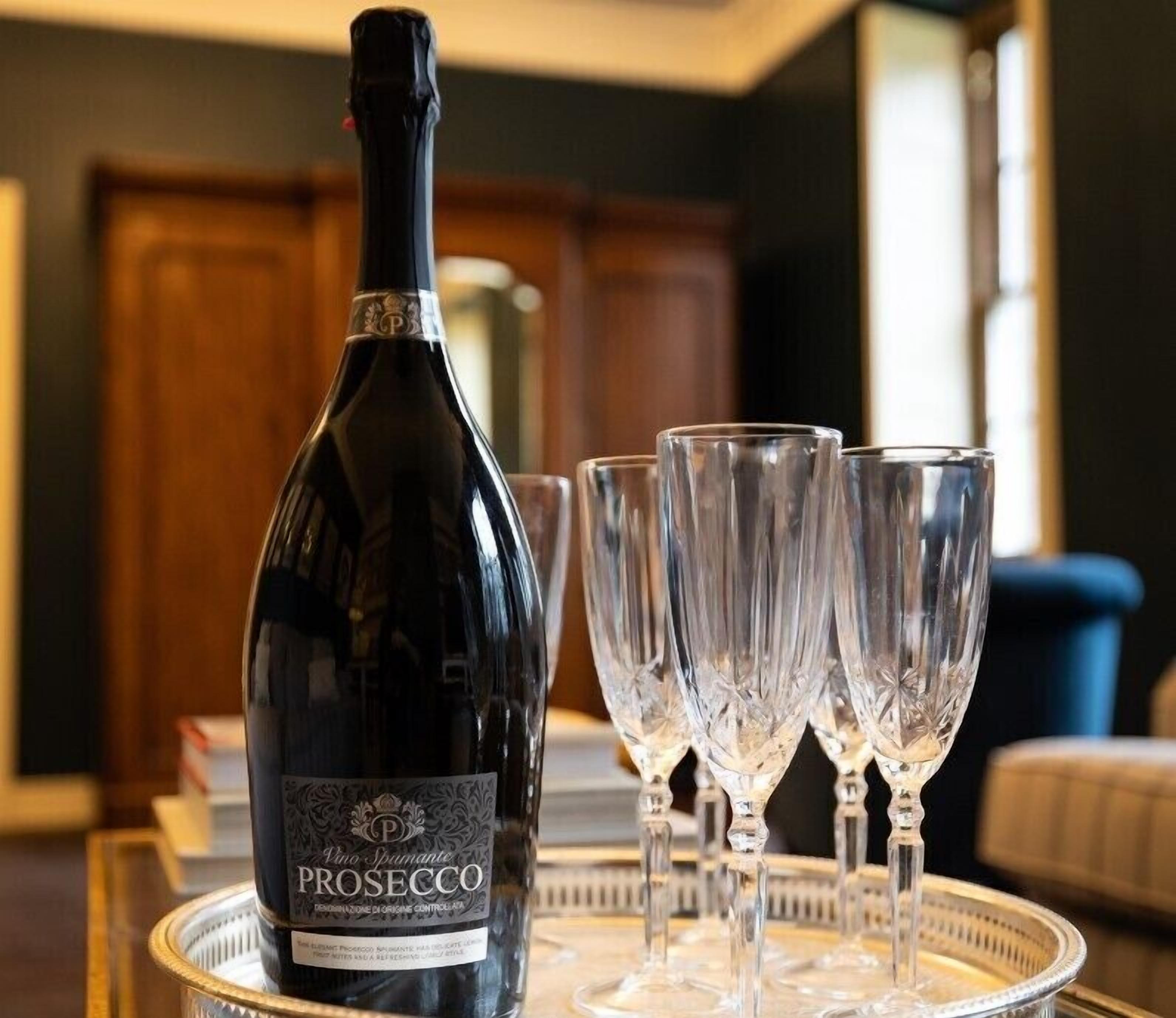 A bottle of Prosecco wine and four crystal wine glasses on a tray in a room with a wooden cabinet and a blue chair in the background.