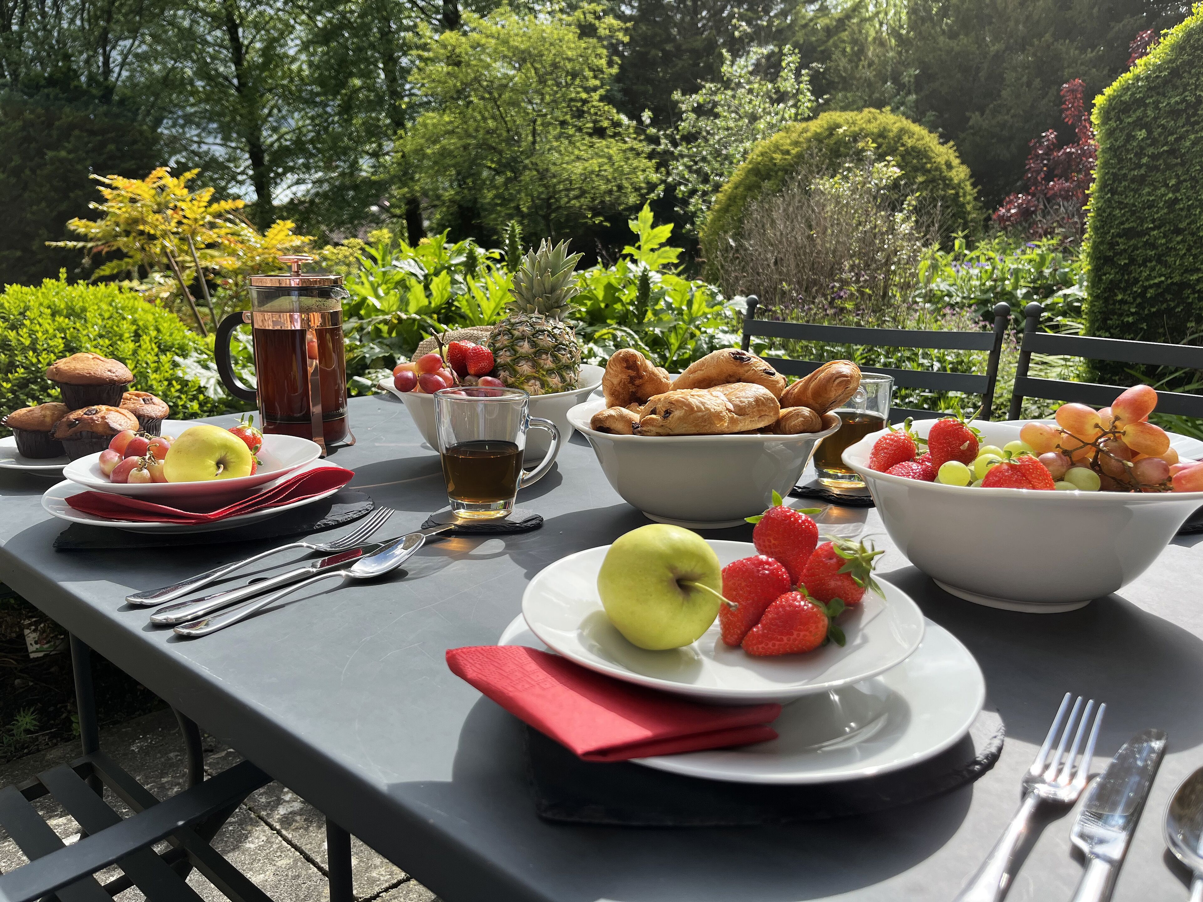 A table set with breakfast items including fruits, pastries, and beverages, surrounded by greenery.