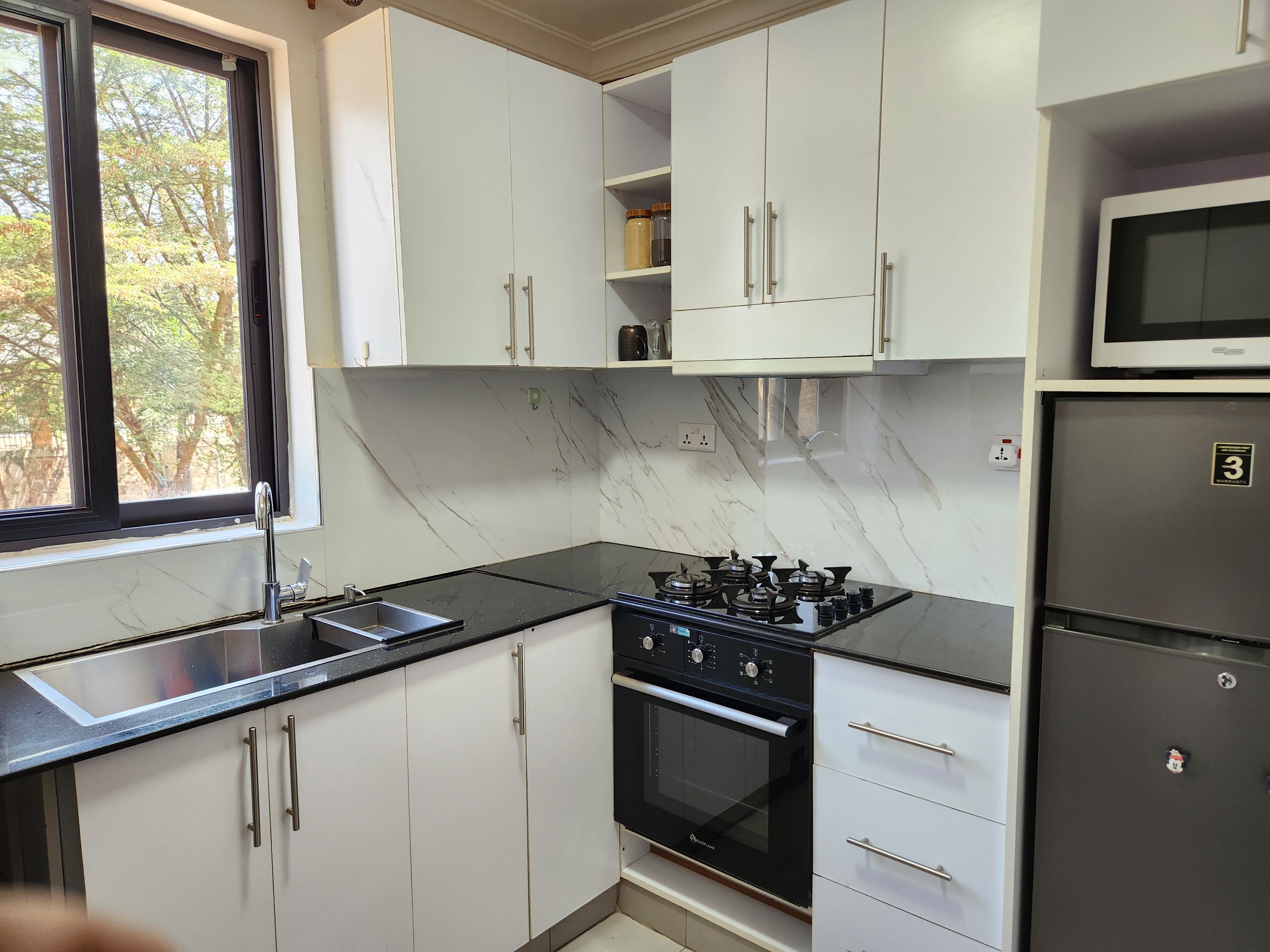 Une cuisine moderne avec des meubles blancs, un plan de travail noir, un réfrigérateur en acier inoxydable et une fenêtre donnant sur des arbres.