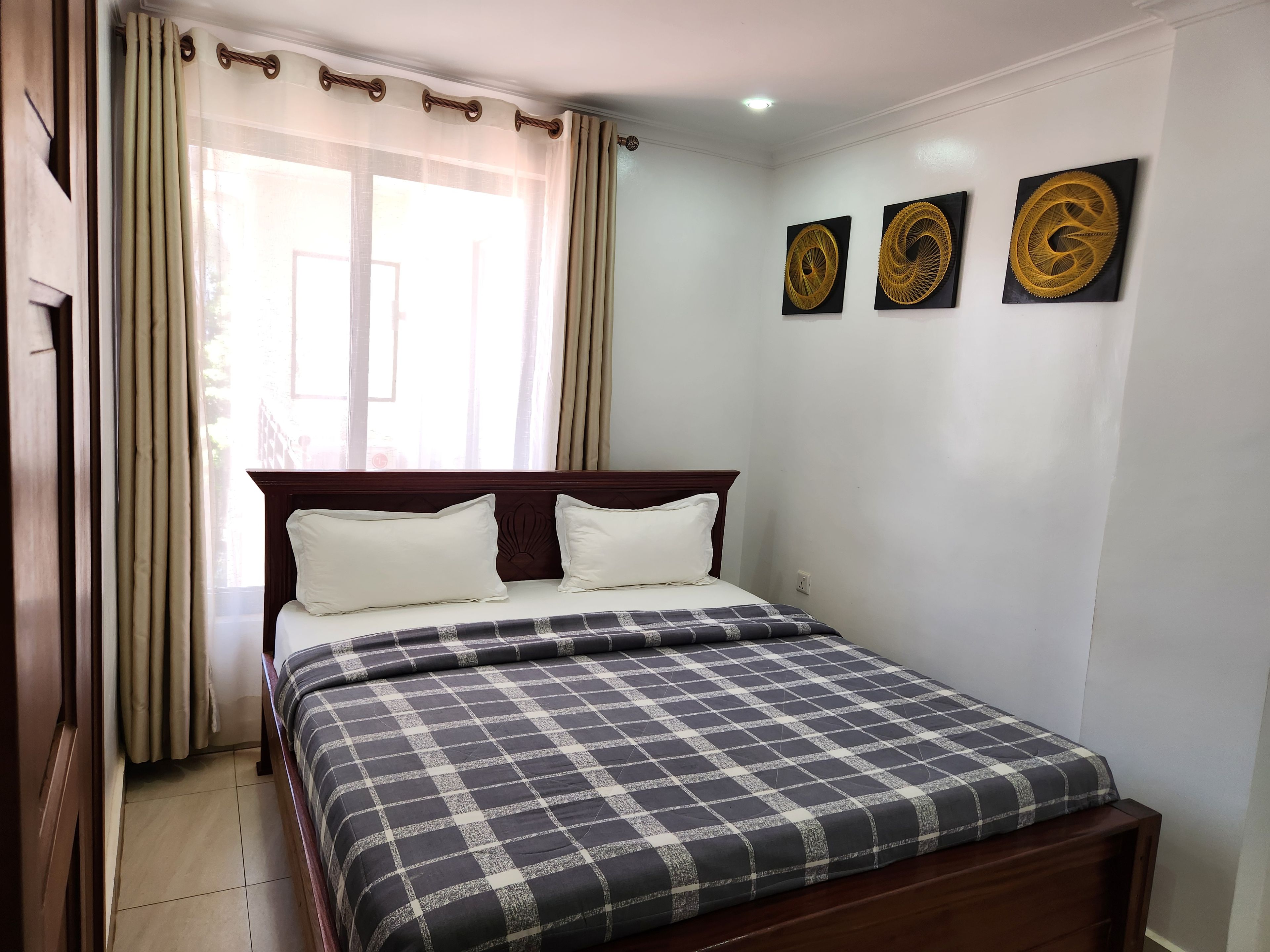 Una cama bien hecha con colcha de cuadros, cabecera de madera y tres obras de arte colgadas en la pared.