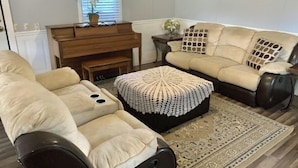 A living room with a beige sofa, a black leather sofa, a coffee table with a crocheted cover, a piano, and a patterned rug.