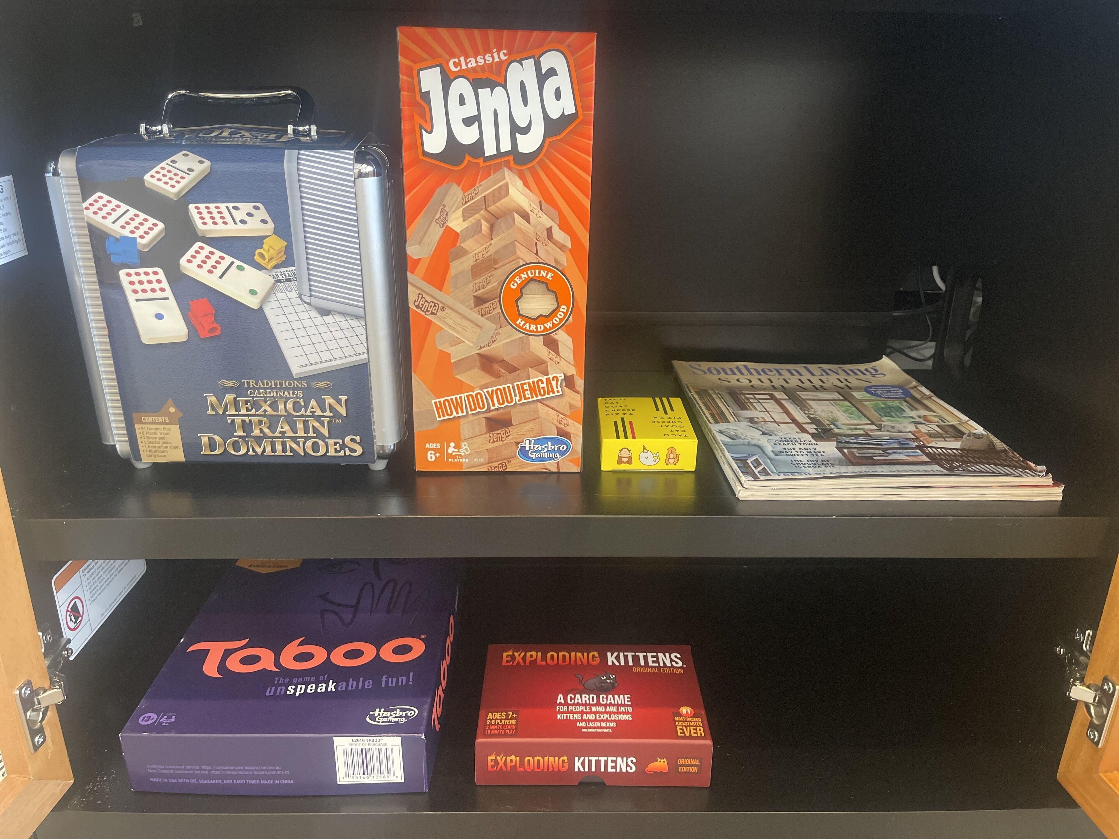 Un estante con varios juegos de mesa, incluyendo 'Mexican Train Dominos', 'Jenga', 'Taboo' y 'Exploding Kittens'.