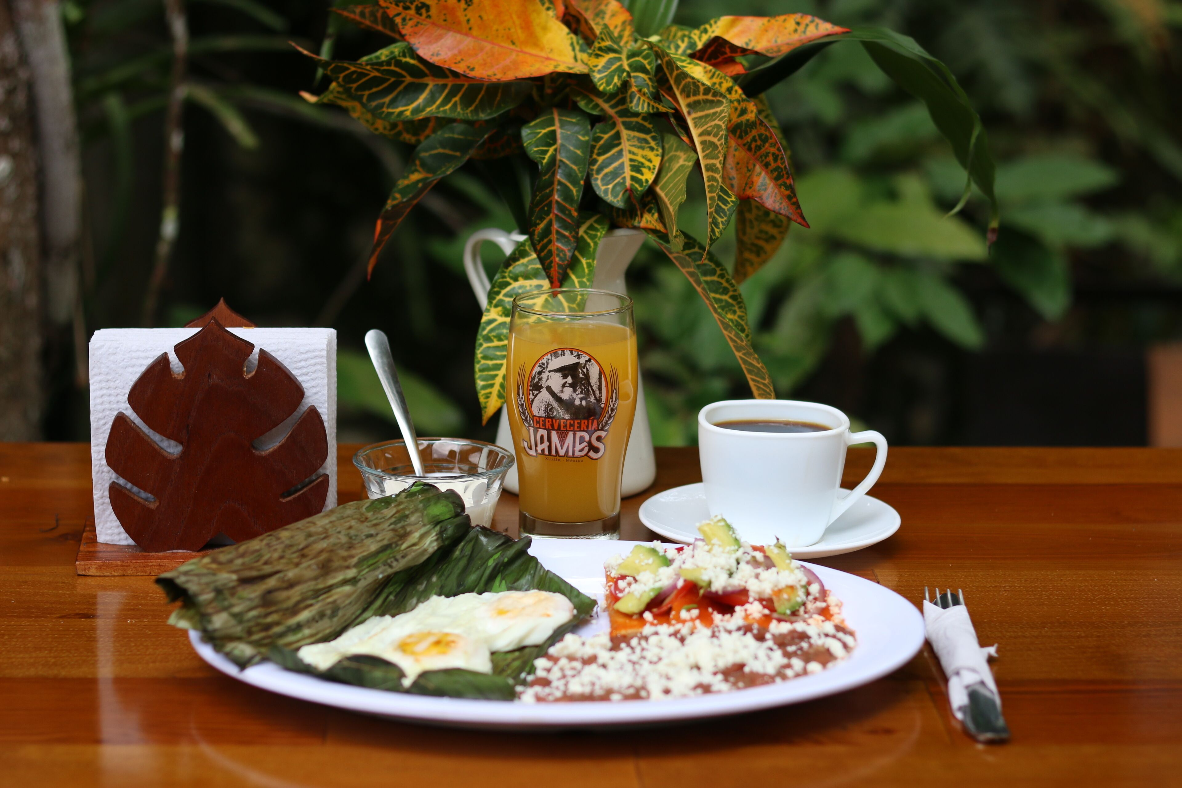 A wooden table with a plate of food, a glass of beer, a cup of coffee, and a napkin.
