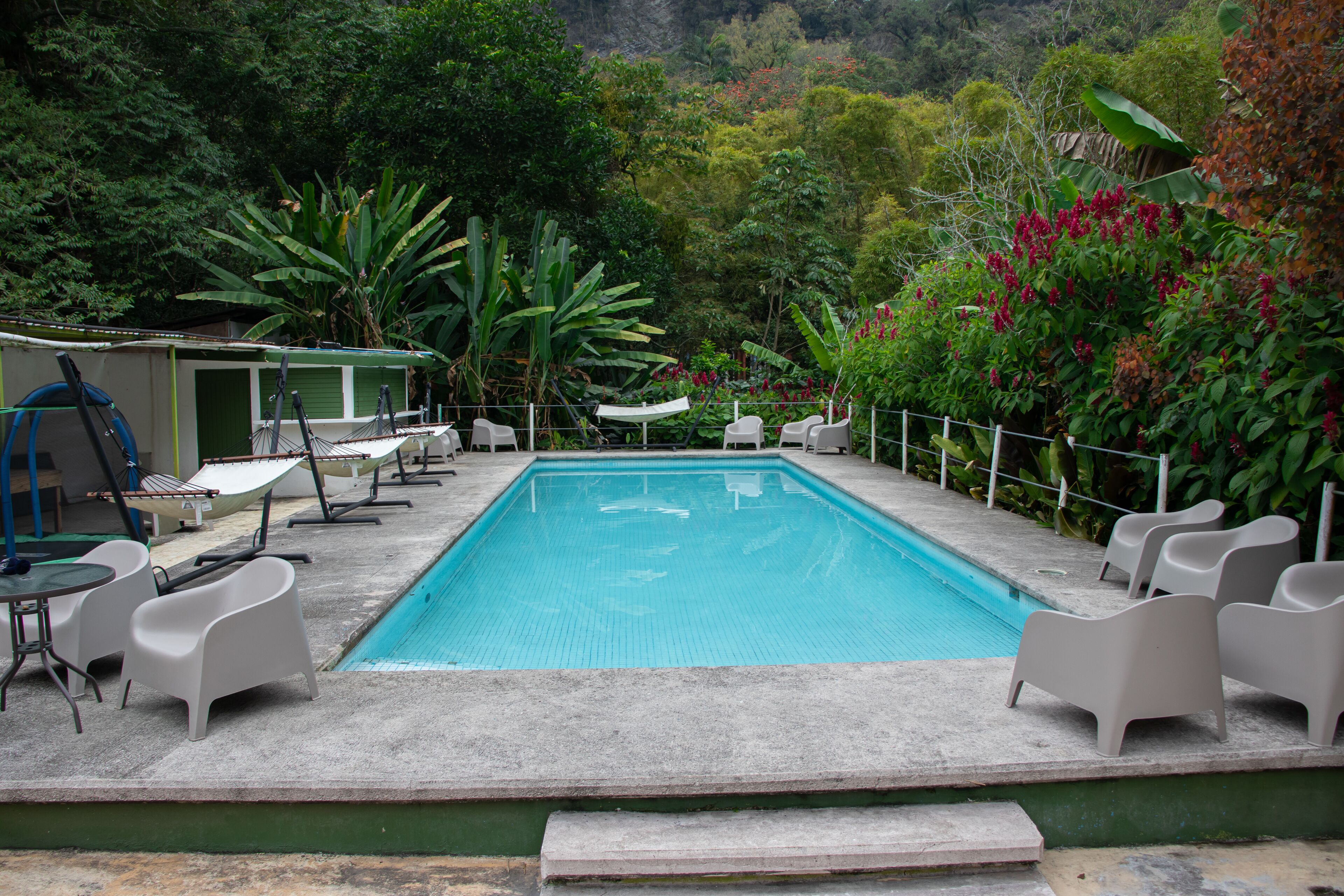 A rectangular swimming pool with a concrete deck, surrounded by white lounge chairs and a small table.