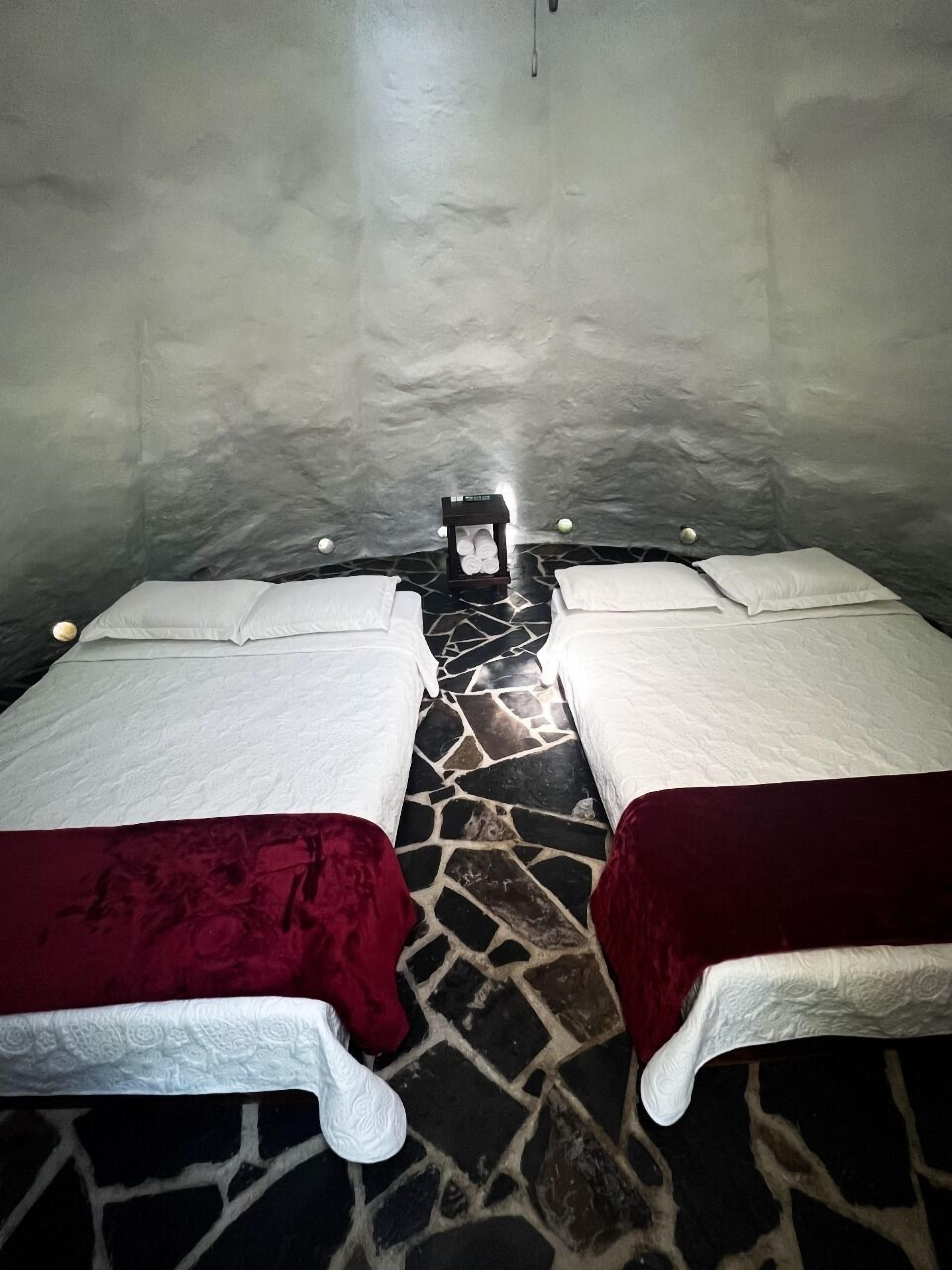 A room with two beds, each with a red blanket and white pillows. There is a small table with a candle in the center. The floor is made of stone tiles.