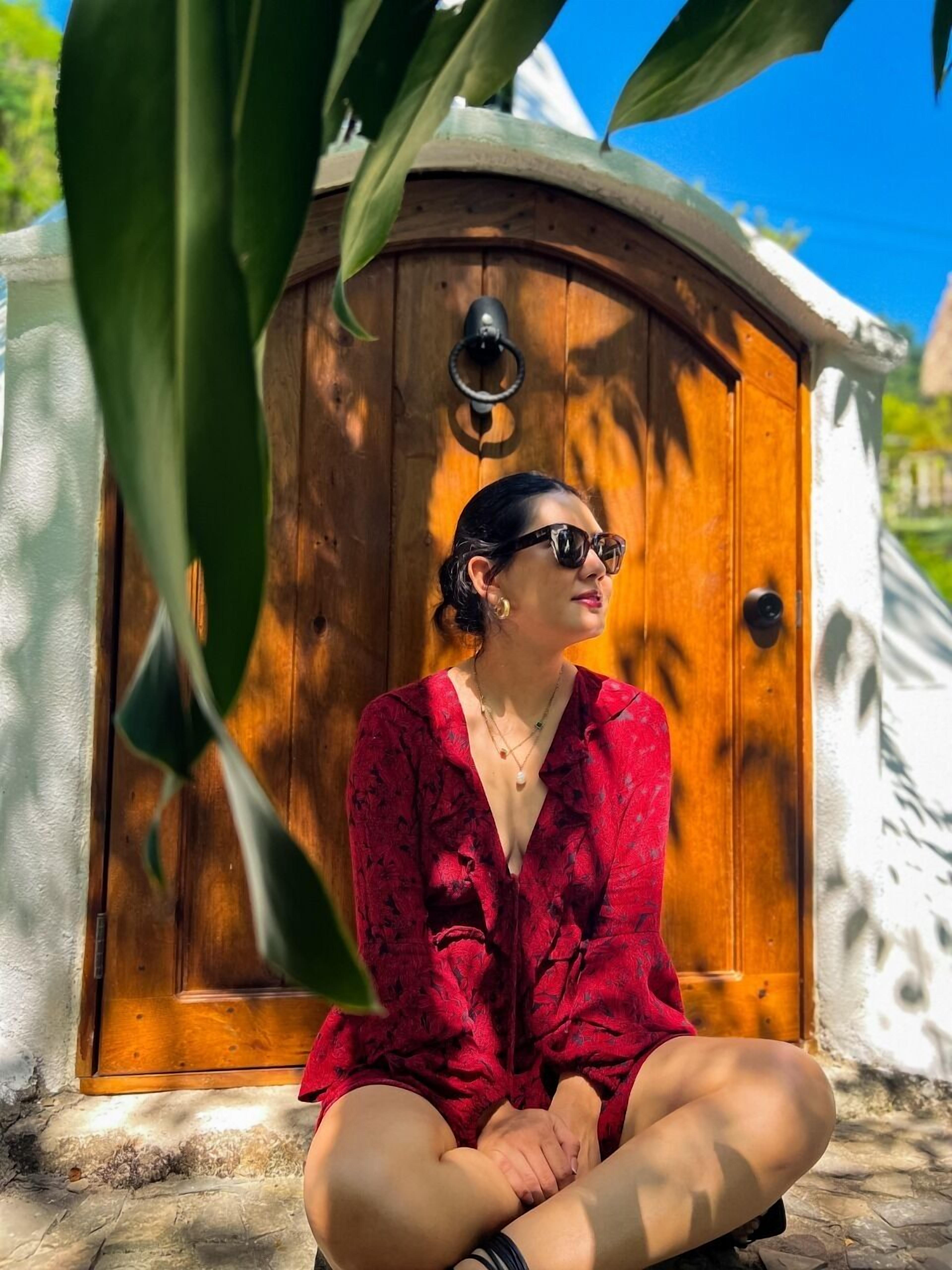 A person sitting in front of a wooden door with a brass knocker, surrounded by greenery.