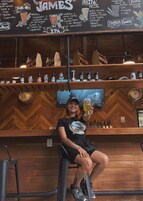 A bar area with a chalkboard menu listing various drinks, a person holding a beer, and shelves with bottles and glasses.