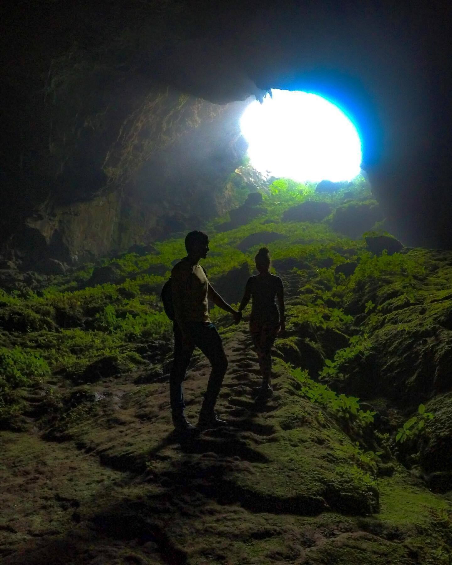 Two people walking on a rocky path inside a cave with a bright light at the end.