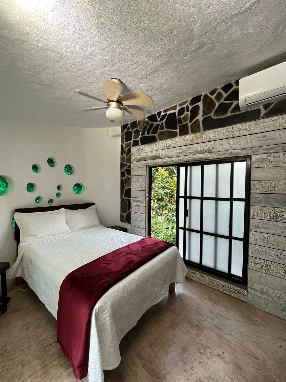 A bedroom with a bed, a ceiling fan, a stone wall, and a window with a view of greenery.