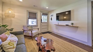 Un salon moderne avec un canapé, une table basse, un téléviseur fixé au mur et une fenêtre équipée de stores.
