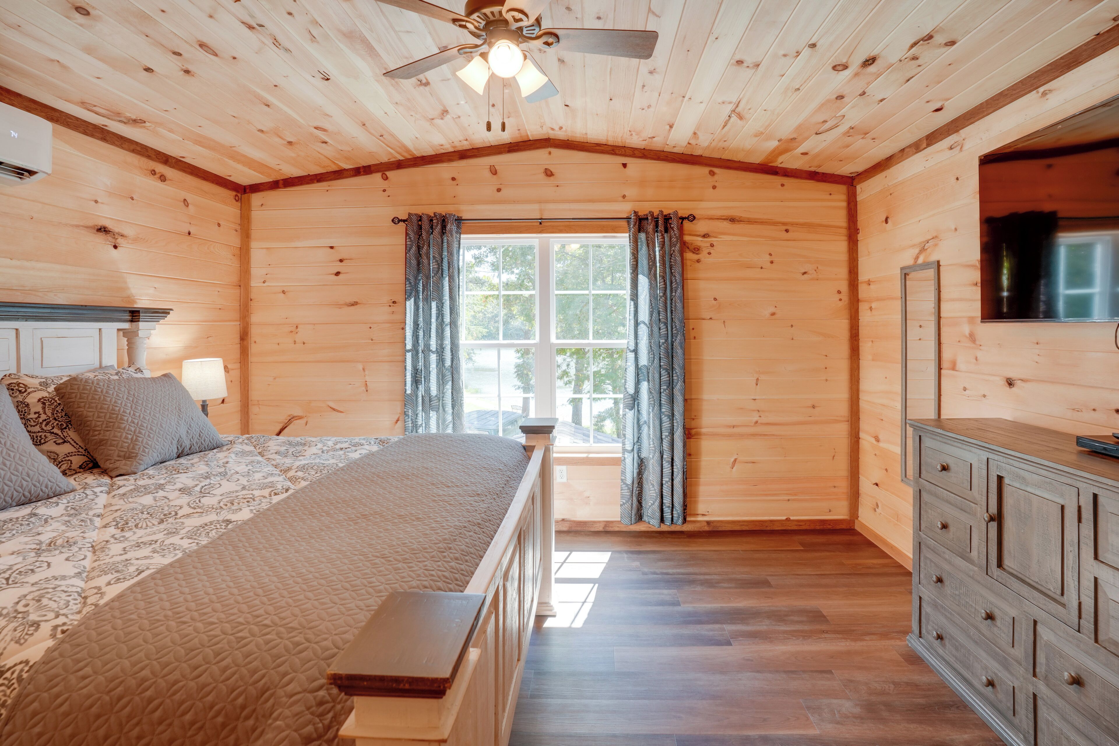 Ein Schlafzimmer einer Holzhütte mit Bett, Kommode und Fenster mit Vorhängen.