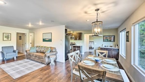 A spacious living room with hardwood flooring, a dining area with a wooden table and chairs, a kitchen with dark cabinets, and a large window providing a view of the outdoors.
