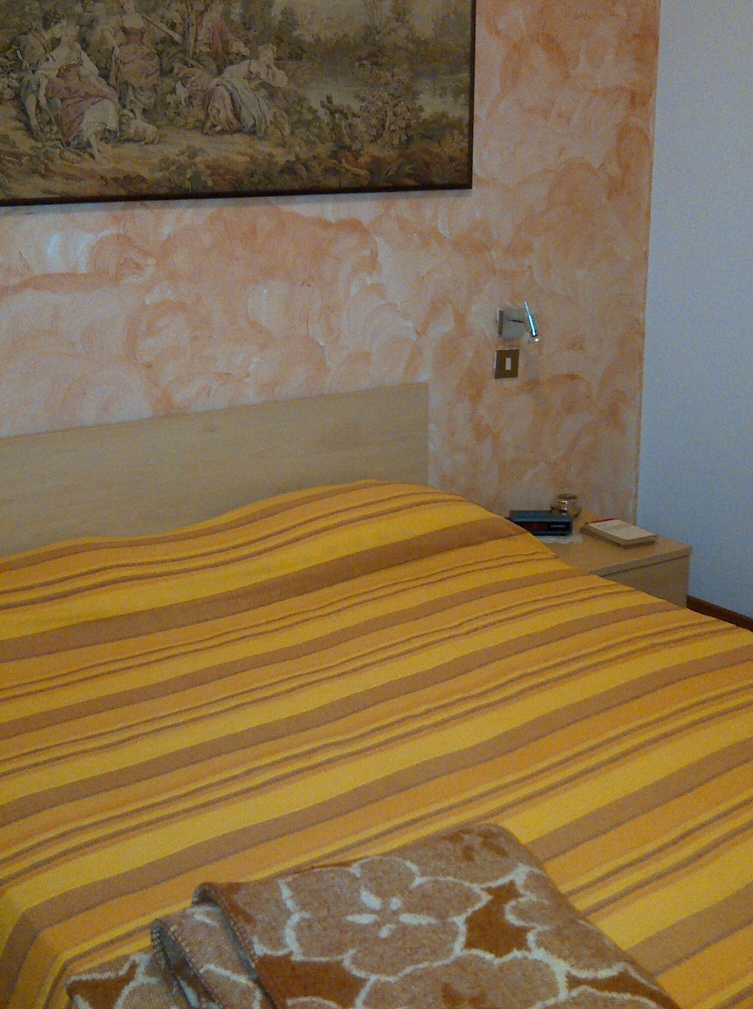 Hotellihuone, jossa on sänky, yöpöytä, taulu seinällä ja termostaatti.