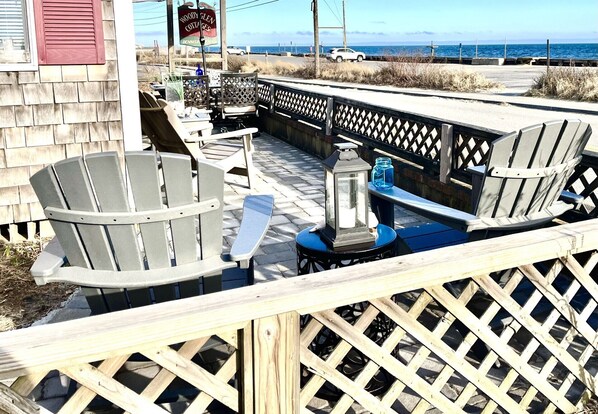 A deck with Adirondack chairs and a table, overlooking a beach and ocean.