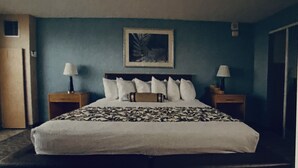 Une chambre d’hôtel avec un grand lit, deux tables de chevet, une petite table, un tableau encadré au mur et une porte sur le côté gauche.