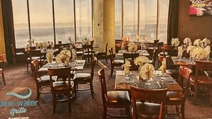 Un restaurant avec des tables dressées pour le service, de grandes fenêtres et une vue sur l’océan.