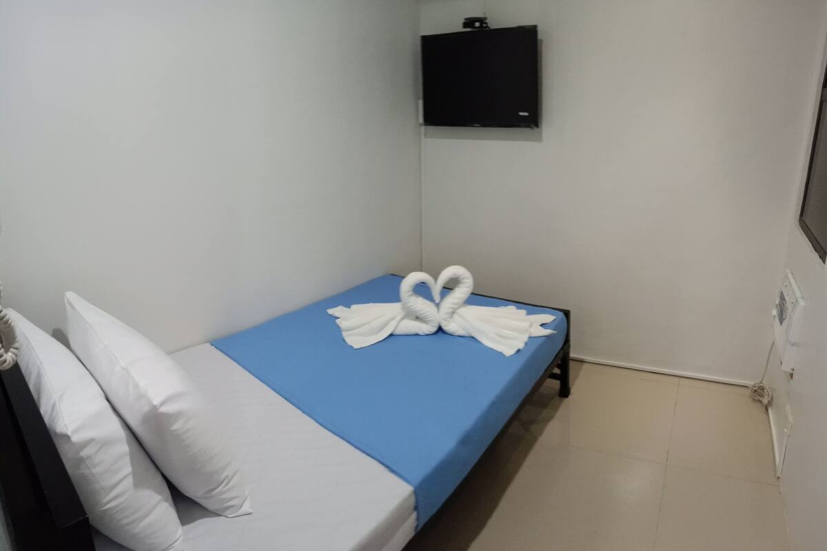 A single bed with a blue mattress cover, white pillows, and a towel arranged in a heart shape.
