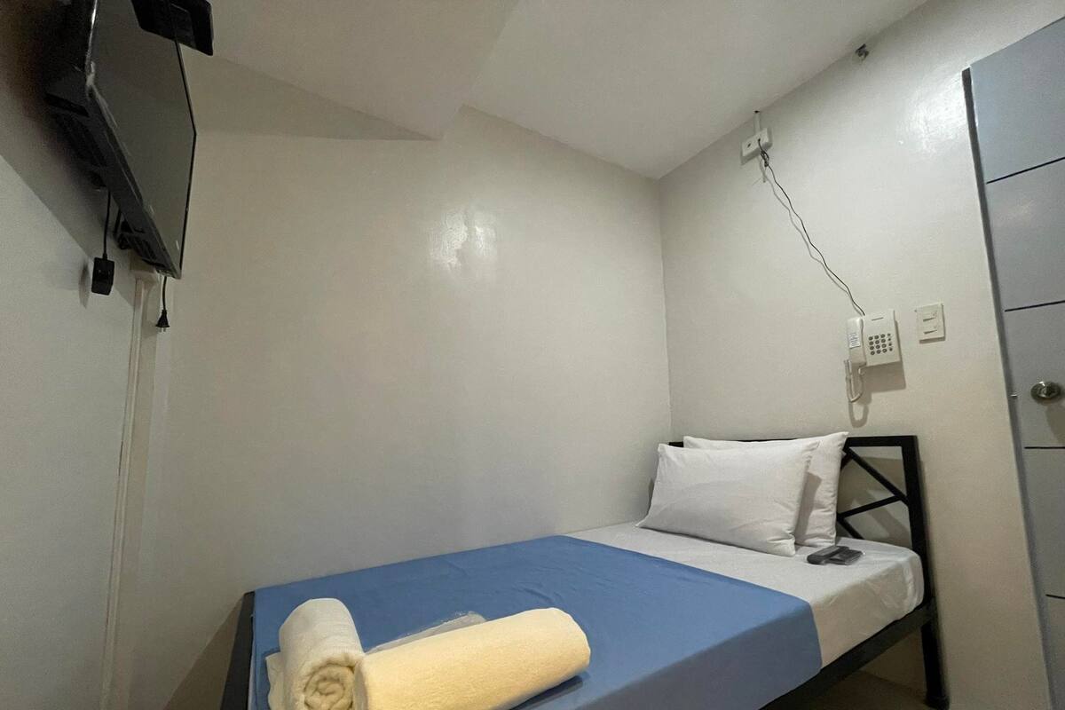 A single bed with a blue mattress and white pillows, a rolled white towel, a bedside telephone, and a TV mounted on the wall.
