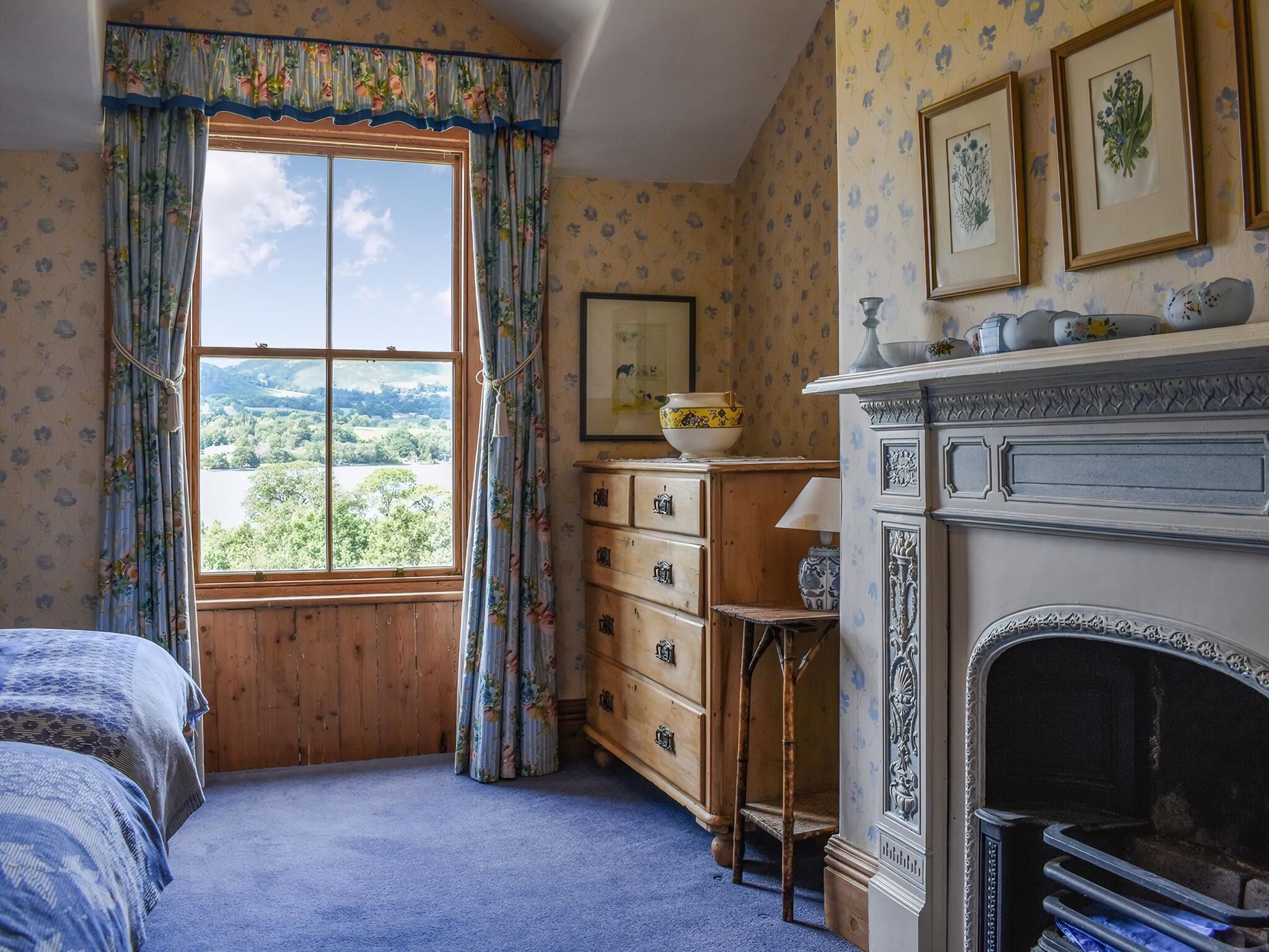 A bedroom with a large window, wooden dresser, and a fireplace.