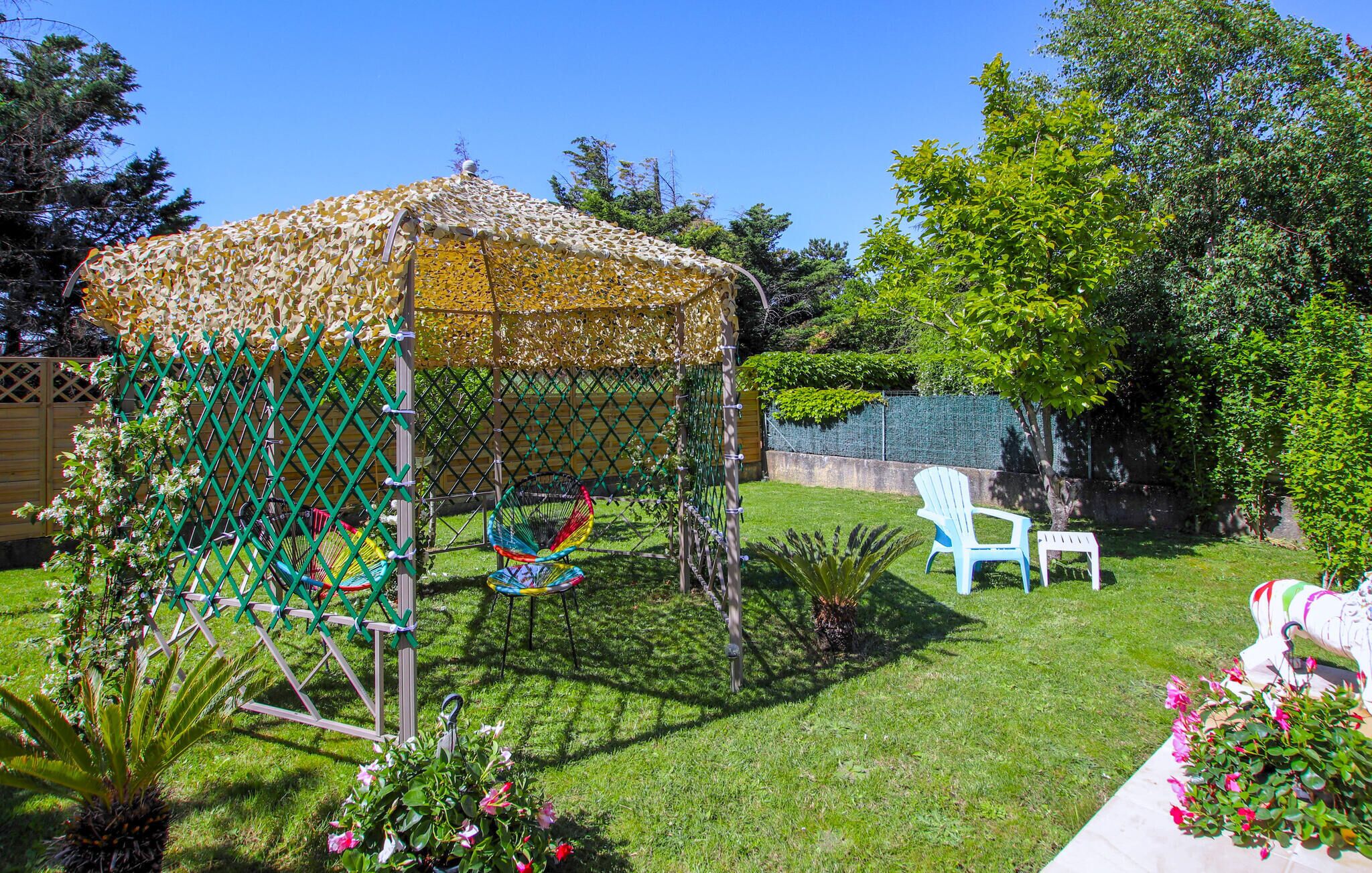 A gazebo with a lattice roof, surrounded by a green fence, and two chairs in the background.