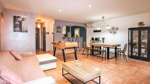 A cozy living room with a sofa, a coffee table, and a foosball table. There is a dining area with a table and chairs, and a kitchen area with a fridge and a sink.