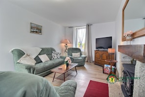 A living room with a sofa, armchair, and a television. There is a fireplace and a window with curtains. - Au Vieux Cupidon - Josselin (JOSSELIN)