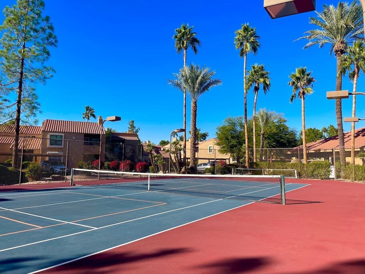 An outdoor tennis court with a net, surrounded by palm trees and buildings.