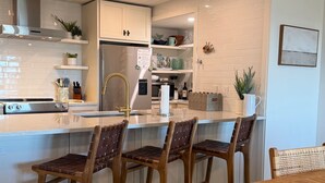 A modern kitchen with white cabinetry, a stainless steel refrigerator, and a wooden dining table with four chairs. - Beachfront w/ Breathtaking Views! 1BR/2BA  Amenity Access included! (Seabrook Island)