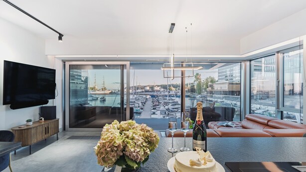 Ein modernes Wohnzimmer mit großem Fenster, Flachbildfernseher, Holzschrank, Esstisch mit Kuchen und Champagner sowie eine Ledersofa.