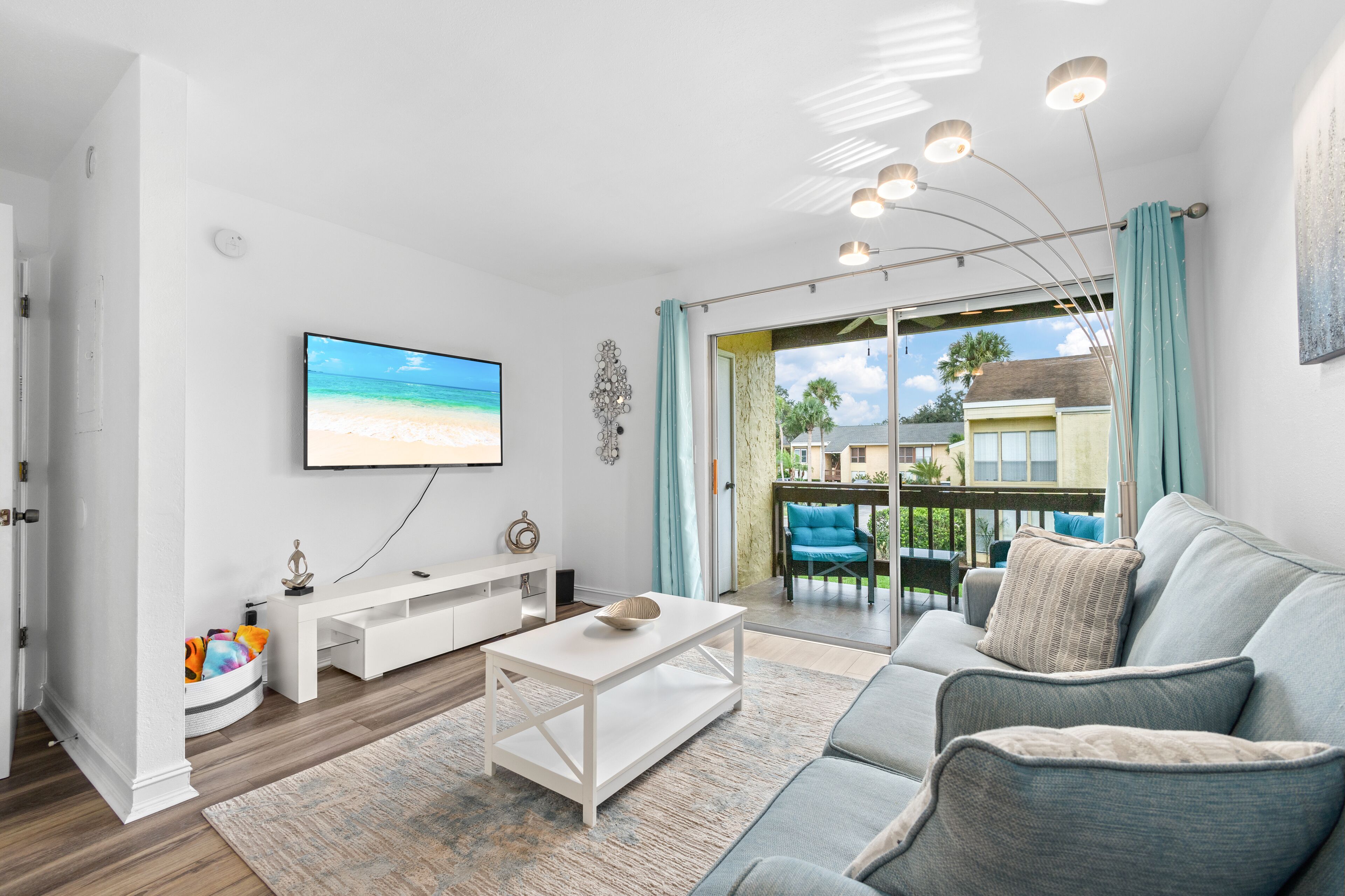 A modern living room with a flat-screen TV, a white coffee table, a grey sofa, and a view of the outdoors through a sliding glass door.