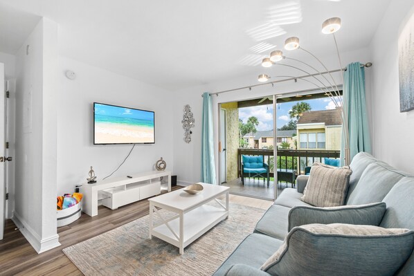 A modern living room with a flat-screen TV, a white coffee table, a grey sofa, and a view of the outdoors through a sliding glass door.