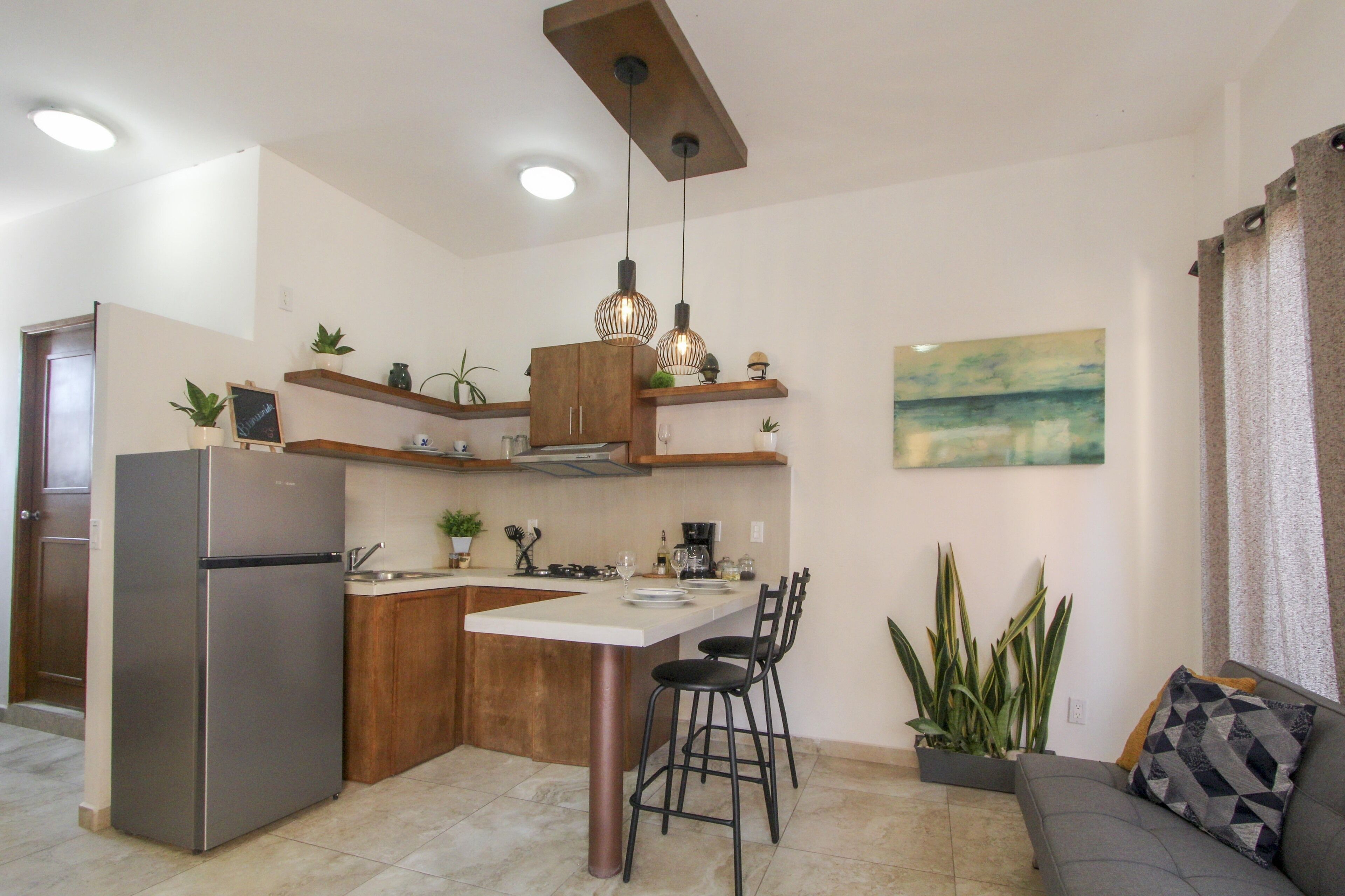 A compact kitchen with a refrigerator, a dining table with chairs, and a coffee machine. There are potted plants and a painting on the wall.