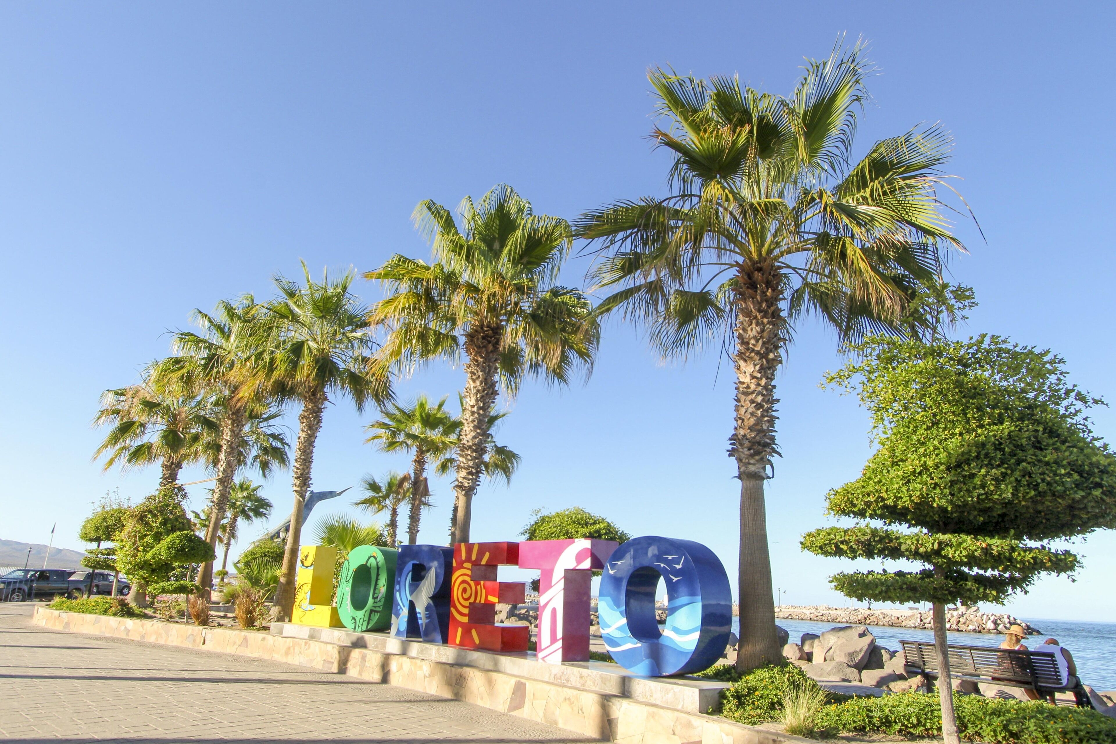 Ein Schild mit dem Wort "ORETTO" in bunten Buchstaben, Palmen und Blick auf das Meer.
