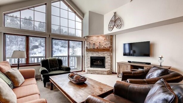 A spacious living room with a stone fireplace, leather sofas, a large TV, and a view of a snowy landscape through large windows.