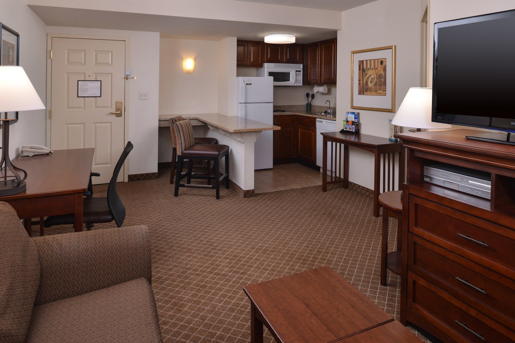 A compact hotel room with a kitchenette, a dining area, a living room with a sofa and TV, and a wooden cabinet.