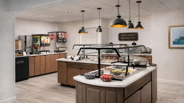 A modern kitchen with a breakfast bar, a Coca-Cola vending machine, and a sign offering free daily breakfast. - Studio | Free Breakfast + Outdoor Pool and Hot Tub (San Antonio)