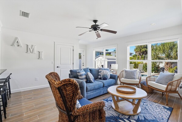 A spacious living room with a blue sofa, wicker chairs, a ceiling fan, and large windows offering a view of the outdoors. - Island Interlude: Roomy Coastal Stay | Heated Pool | Near Beach (Holmes Beach)